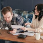 A couple sitting at a desk, reviewing bills and expenses with a calculator and laptop, looking worried.