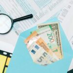 Close-up of tax documents and mixed currency in a blue envelope with a magnifying glass, perfect for financial analysis concepts.