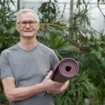 Smiling senior man with gray hair holding a yoga mat in a lush greenhouse.