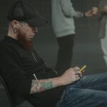 A tattooed man in casual attire using a smartphone at an indoor social event with other attendees.