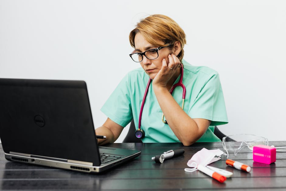 Focused nurse with eyeglasses working on a laptop surrounded by medical tools on a desk.