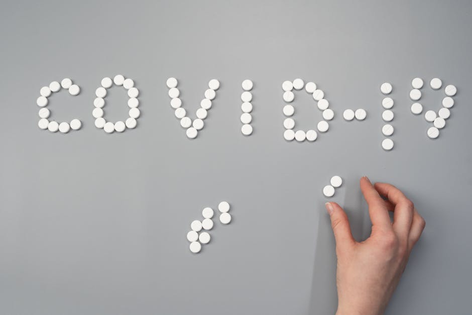 Hand arranging pills spelling 'COVID-19' on a gray background, depicting healthcare theme.